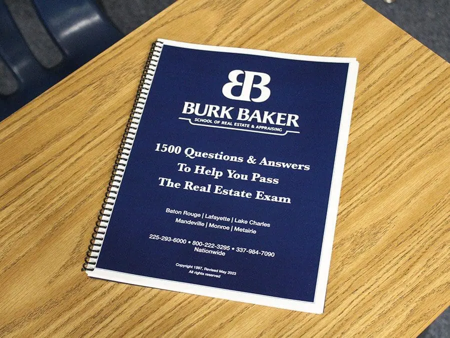 Blue spiral-bound book titled "Burk Baker School of Real Estate & Appraising" placed on a wooden desk. The cover text reads "1500 Questions & Answers To Help You Pass The Real Estate Exam" followed by the locations: Baton Rouge, Lafayette, Lake Charles, Mandeville, Monroe, Metairie, and contact numbers.