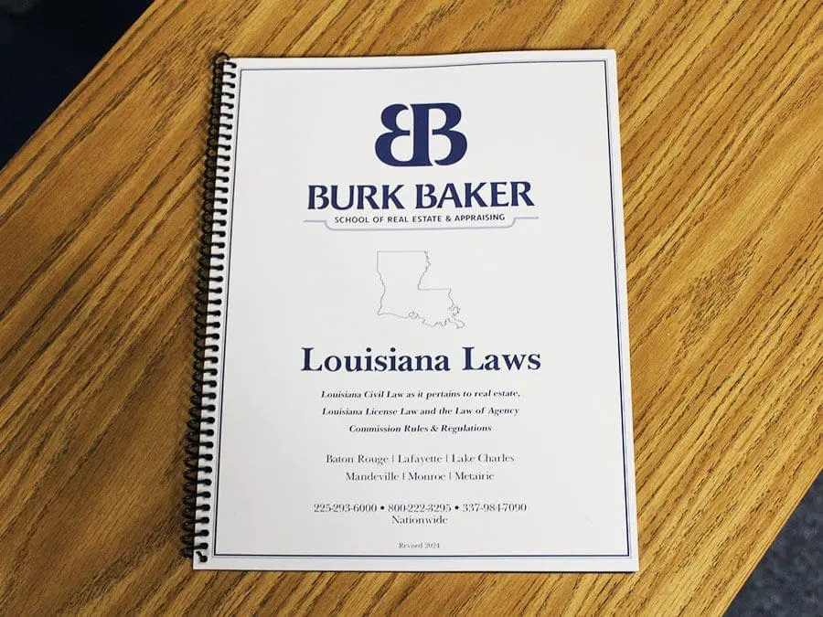 A spiral-bound book titled "Louisiana Laws" is on a wooden table. The cover features the logo "BB" and the text "BURK BAKER SCHOOL OF REAL ESTATE & APPRAISING." There is an outline of the state of Louisiana. The book includes information about Louisiana Civil Law, License Law, and agency laws. Contact numbers and locations such as Baton Rouge, Lafayette, and others are listed at the bottom.