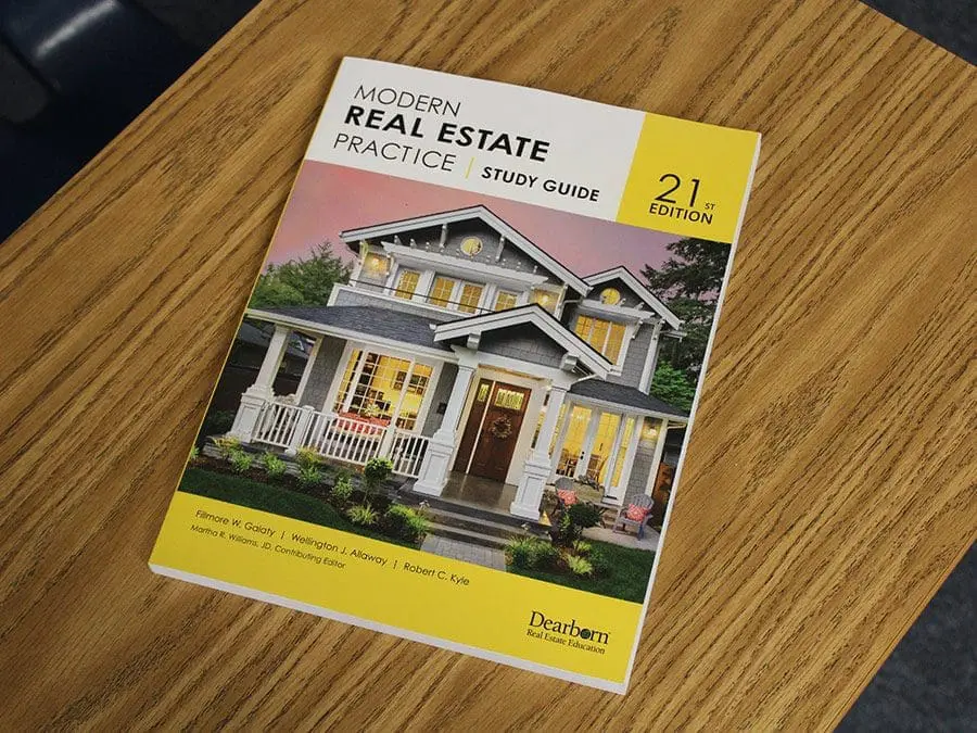 Cover of the "Modern Real Estate Practice" study guide, 21st edition, lying on a wooden table. The cover features an image of a house with a front porch and lights on, surrounded by shrubs and trees. The title and edition number are prominently displayed in black and yellow text. The publisher, Dearborn Real Estate Education, is mentioned at the bottom.