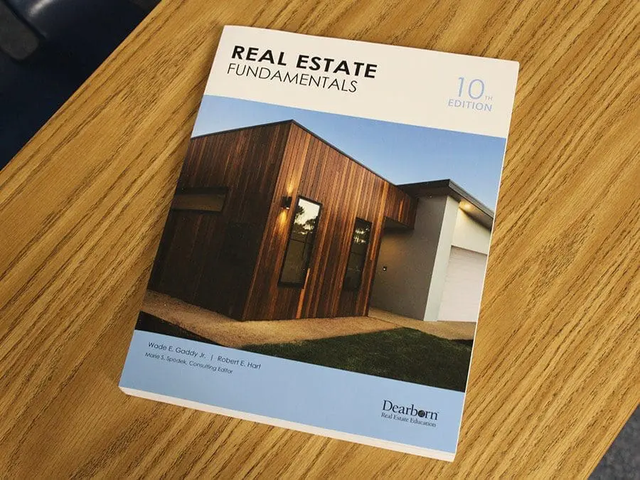Cover of the "Real Estate Fundamentals" textbook, 10th edition, placed on a wooden table. The cover features an image of a modern house with wooden and white exterior elements. The book is published by Dearborn Real Estate Education.