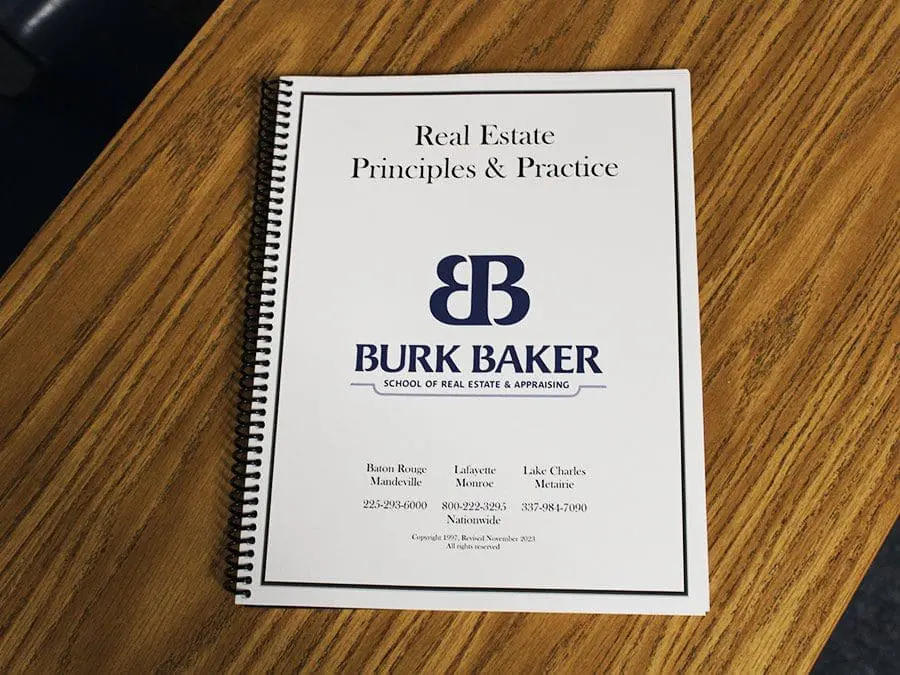 Cover of a spiral-bound book titled "Real Estate Principles & Practice" from Burk Baker School of Real Estate & Appraising. It rests on a wooden surface. Contact information for various locations is printed at the bottom.